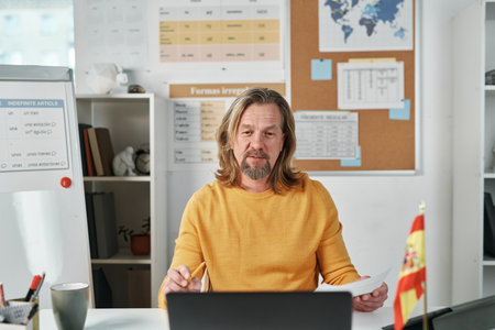 Mature professional teacher sitting at table in front of laptop and talking to students online in classの写真素材