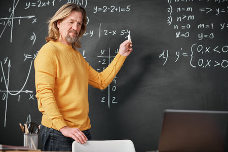 Mature teacher standing near the blackboard and explaining Maths to students online using laptopの写真素材