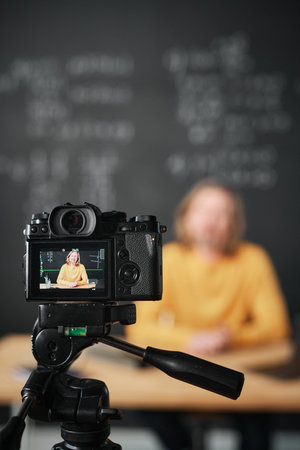 Vertical image of teacher sitting at table against blackboard and recording lesson on professional...