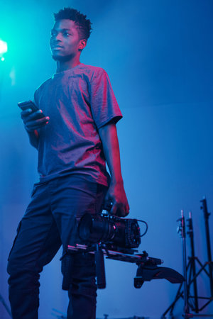 African American video operator standing in dark studio with smartphone and professional cameraの写真素材