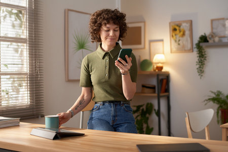 Young woman sitting in the kitchen enjoying her coffee and reading message on her smartphoneの写真素材