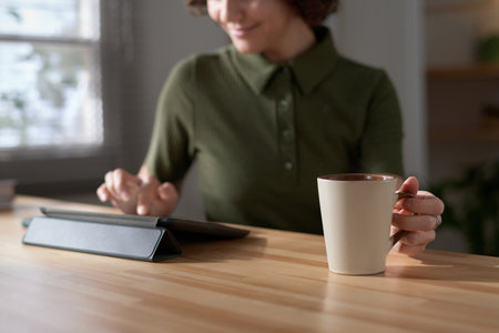 Close-up of young woman sitting at kitchen table drinking coffee and typing on digital tabletの写真素材
