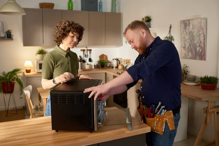 Repairman using his tools to fix microwave oven while standing in the kitchen together with housewifeの写真素材