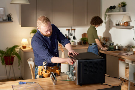 Mature handyman using his work tools to repair oven at home with housewife doing housework in backgroundの写真素材