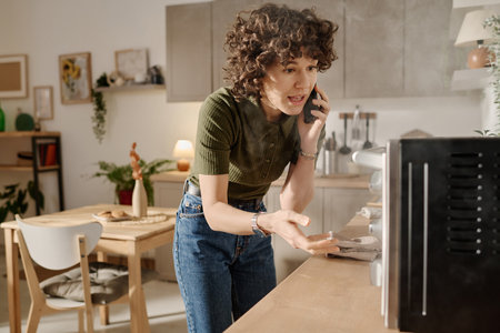 Young woman calling the customer service on breakdown of the oven standing in the kitchen and looking at smoking applianceの写真素材