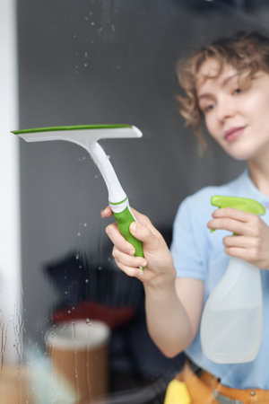 Vertical image of young professional worker cleaning the window with detergent and tools at home