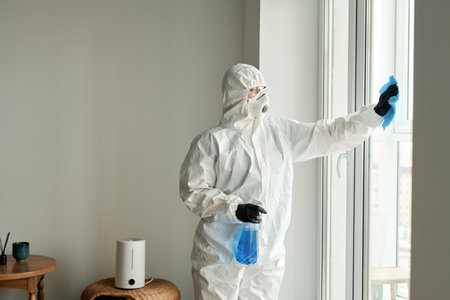 Worker in protective suit wiping windows with rag using detergent during housework in apartmentの写真素材