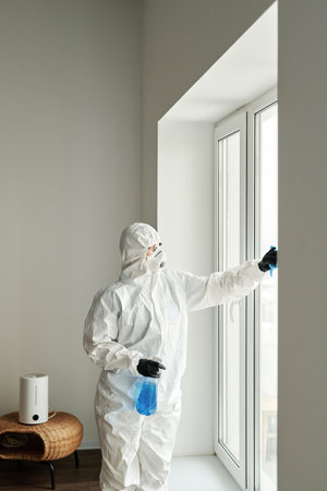 Vertical image of cleaning worker in protective suit wiping windows with detergent while working in apartmentの写真素材