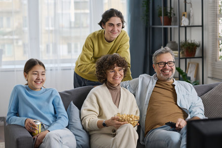 Happy family of four sitting on sofa with popcorn and watching movie on TV together in the living roomの写真素材