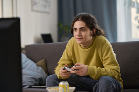 Teenage boy playing game console while sitting on sofa in front of TV screen at home