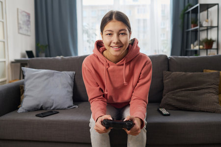 Portrait of smiling teenage girl looking at camera while sitting on sofa in the room and playing video gameの写真素材