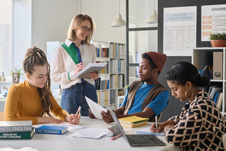 Young smiling teacher conducting foreign language lesson with students while they sitting at table with books and computerの写真素材
