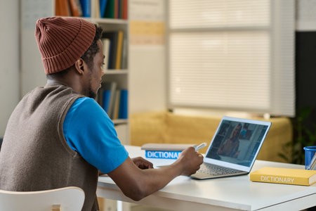 Rear view of African American student having online lesson with English teacher on laptop while sitting at table in classの写真素材