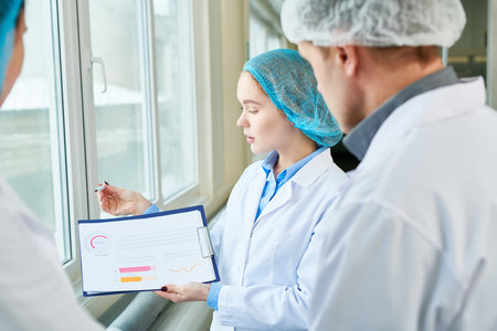 Serious confident female food technologist holding chart and presenting statistic to colleagues while they discussing consumption issues in factoryの写真素材