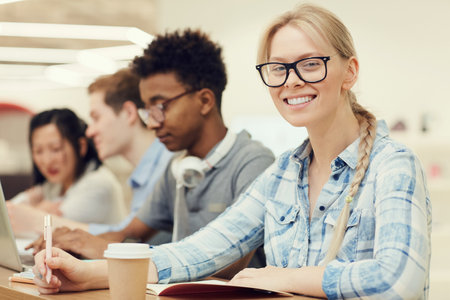 Portrait of cheerful excited student girl with blond braid wearing spectacles sitting at table with groupmates and making notes in workbook at lectureの写真素材