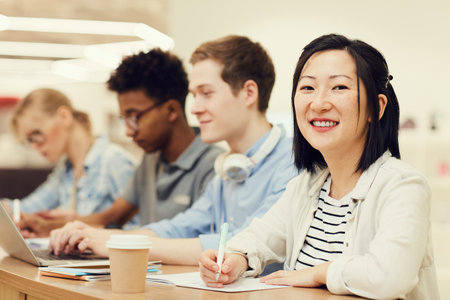 Portrait of smiling young Asian female student with short hair sitting at table with groupmates in lecture room and summarizing informationの写真素材