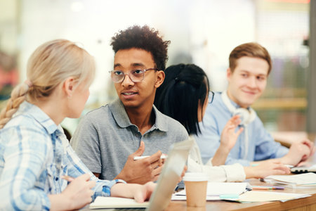 Group of young interracial students sitting at desk with workbooks and devices and discussing scientific projectの写真素材