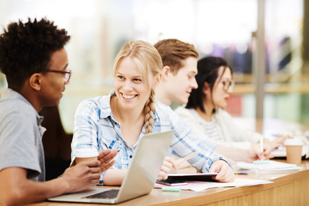 Group of positive multi-ethnic students in casual outfits sitring at one table and chatting in lecture roomの写真素材