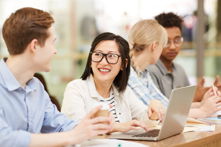 Group of cheerful young interracial students sitting at desk in library and discussing ideas while working on educational projectsの写真素材