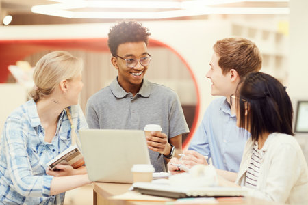 Group of happy young interracial students in casual outfits standing at high table with laptop in library and sharing project ideasの写真素材
