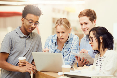 Group of pensive young multi-ethnic students standing at table using wi-fi in library while searching for information on laptopの写真素材