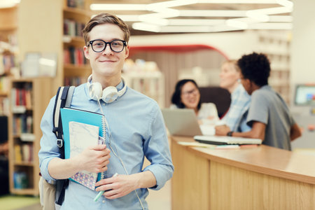 Portrait of smiling handsome young student in spectacles standing in public library and holding workbooksの写真素材