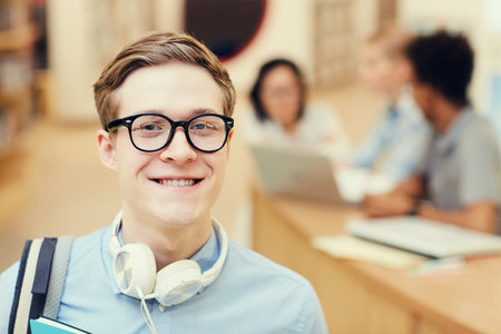 Portrait of smiling smart student boy in eyeglasses wearing headphones on neck spending time in libraryの写真素材