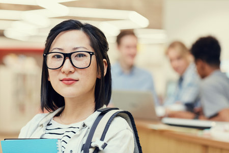Portrait of confident intelligent Asian student girl in glasses wearing satchel standing in university hall, her groupmates chatting in backgroundの写真素材