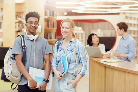 Cheerful multi-ethnic teenage students in casual outfits standing in school library and holding workbooks while posing for cameraの写真素材