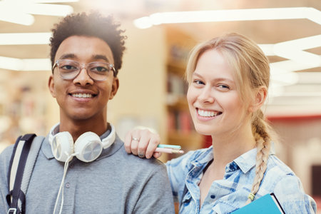 Portrait of positive young interracial students standing in library: pretty girl holding hand on shoulder of Afro-American friendの写真素材