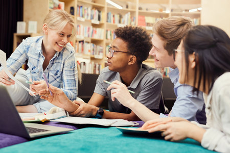 Group of cheerful young multi-ethnic students in casual clothing using laptop and workbooks while doing homework together in libraryの写真素材