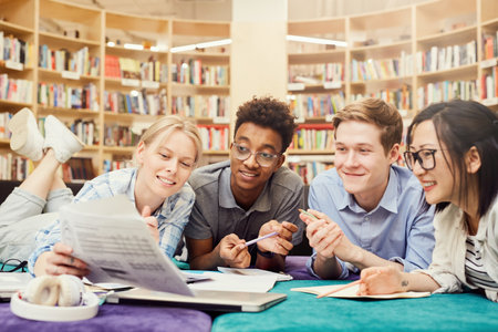 Group of jolly interracial students lying on floor in library and taking training tests while preparing for exam togetherの写真素材