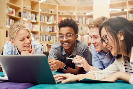 Positive excited multi-ethnic students in casual clothing lying on floor in campus library and laughing while watching curious video on laptopの写真素材