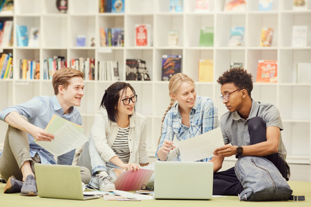 Group of content confident young multi-ethnic students in casual outfits sitting on floor in library and discussing task togetherの写真素材