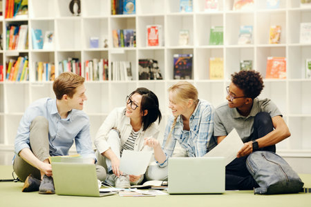 Group of excited young multi-ethnic students sitting on floor in campus library and working on research projectの写真素材