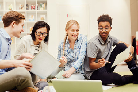 Group of cheerful interracial university students in casual outfits sitting on floor in library and working with paper and digital sources togetherの写真素材