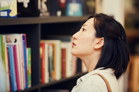 Close-up of serious pensive young Asian woman with short hair standing in bookstore and choosing book on shelfの写真素材
