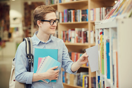 Smiling handsome student boy in eyeglasses standing at shelf and choosing textbook while buying it in bookstoreの写真素材