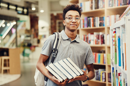 Portrait of content confident African teenage boy in spectacles standing in library and choosing textbooks to do research projectの写真素材