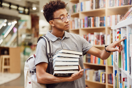 Serious Afro-American high school student in eyeglasses wearing satchel on one shoulder standing in modern library and finding booksの写真素材