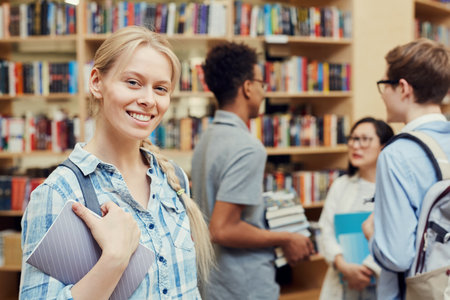 Portrait of cheerful pretty girl with blond hair standing in high school library and holding notepad, her classmates discussing books in backgroundの写真素材