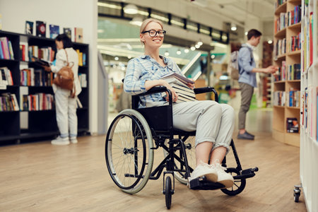 Portrait of happy smart person with disability girl with blond hair sitting in wheelchair and holding books in university libraryの写真素材
