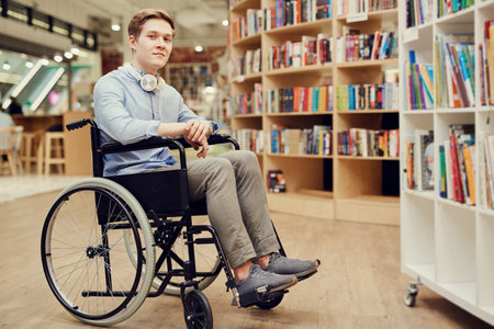Portrait of content student boy with paralyzed legs sitting in wheelchair and choosing books in libraryの写真素材