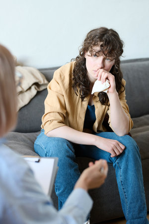 Young woman with handkerchief sitting on sofa and listening to advice of therapist during sessionの写真素材