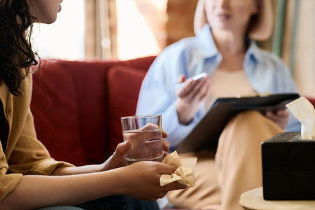 Close-up of young woman with handkerchief and glass of water talking about her problems to psychologistの写真素材