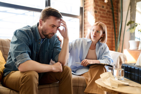 Man listening to advice of psychotherapist while sitting on sofa during his visit to psychologist officeの写真素材