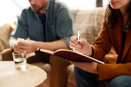 Close-up of young psychologist making notes during conversation with man at sessionの写真素材