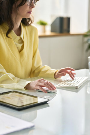 Vertical image of young businesswoman sitting at her workplace in front of computer monitor and typing on keyboardの写真素材