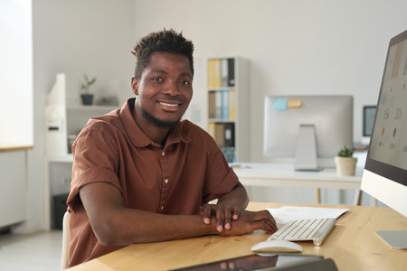 Portrait of African American young businessman smiling at camera while sitting at his workplace in officeの写真素材