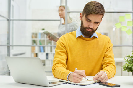 Serious busy handsome young businessman in yellow sweater writing ideas in diary while working in modern officeの写真素材